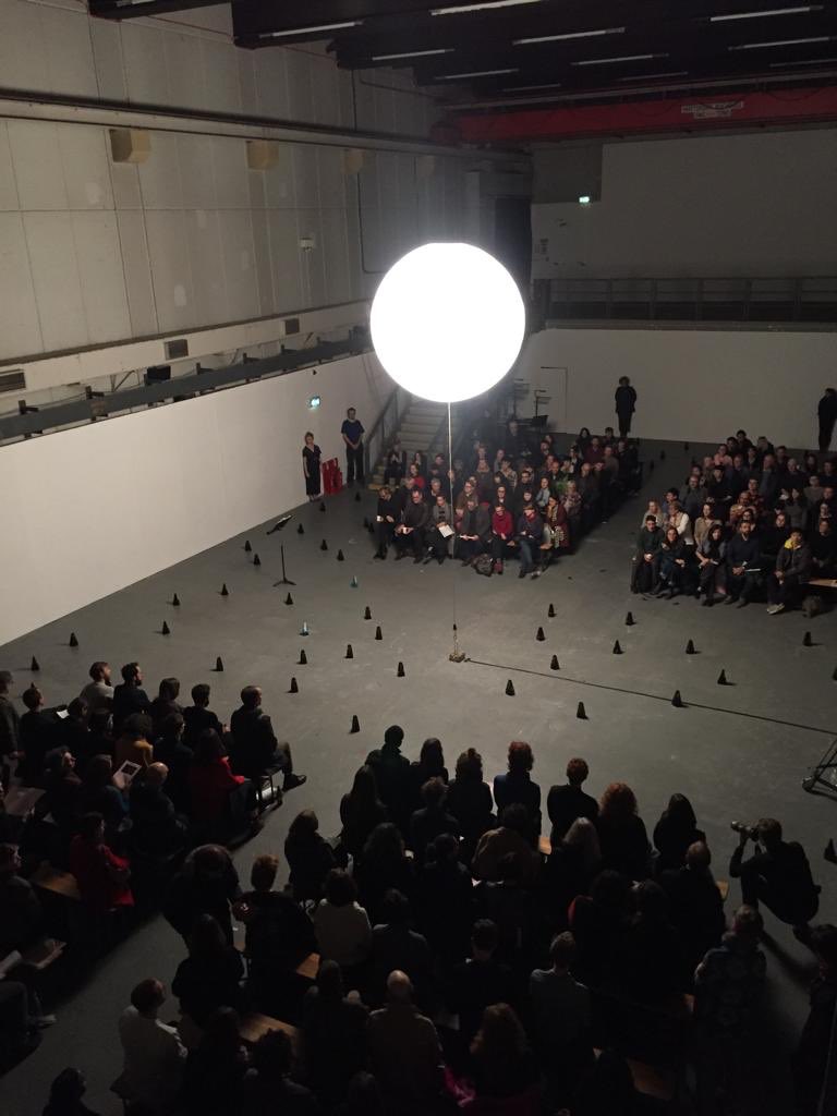 A glowing orb suspended over an audience and an assortment of metronomes in a darkened room