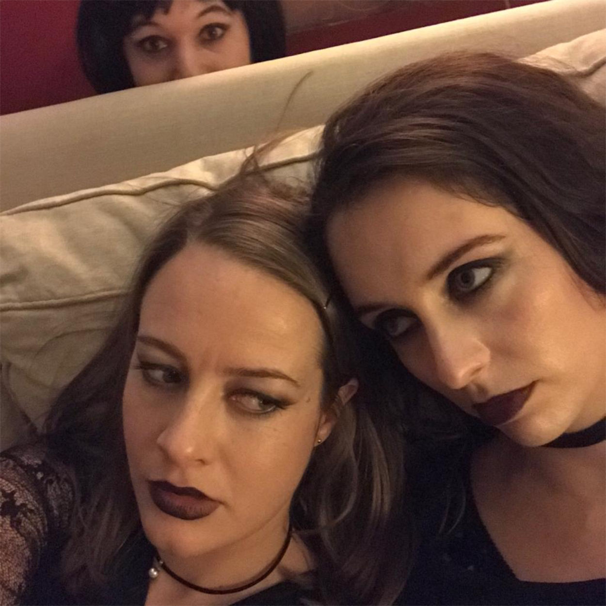 Two girls with dark makeup on sat on a couch with someone peeking up behind
