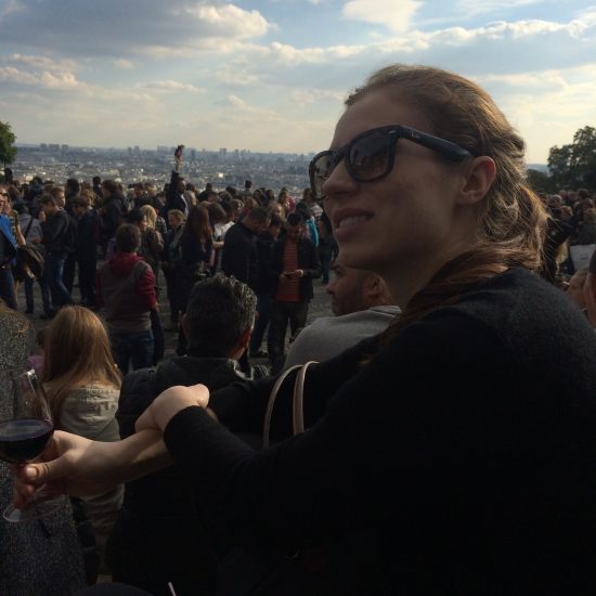A woman with sunglasses smiling with a glass of wine on the steps of the Sacré-Cœur in Paris