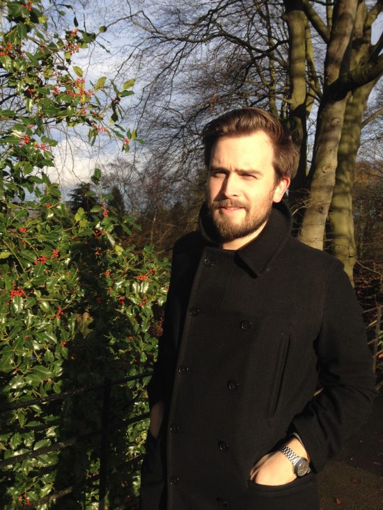 A man in a black pea coat standing in front of holly bushes as the sun sets