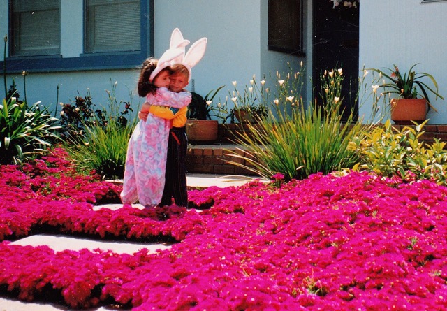 Two kids hugging in bunny ears in front of a light blue house amongst pink ice plant