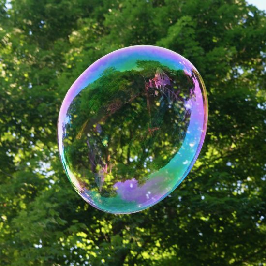 A large, iridescent bubble with a faint reflection of a house floating in front of a leafy green tree