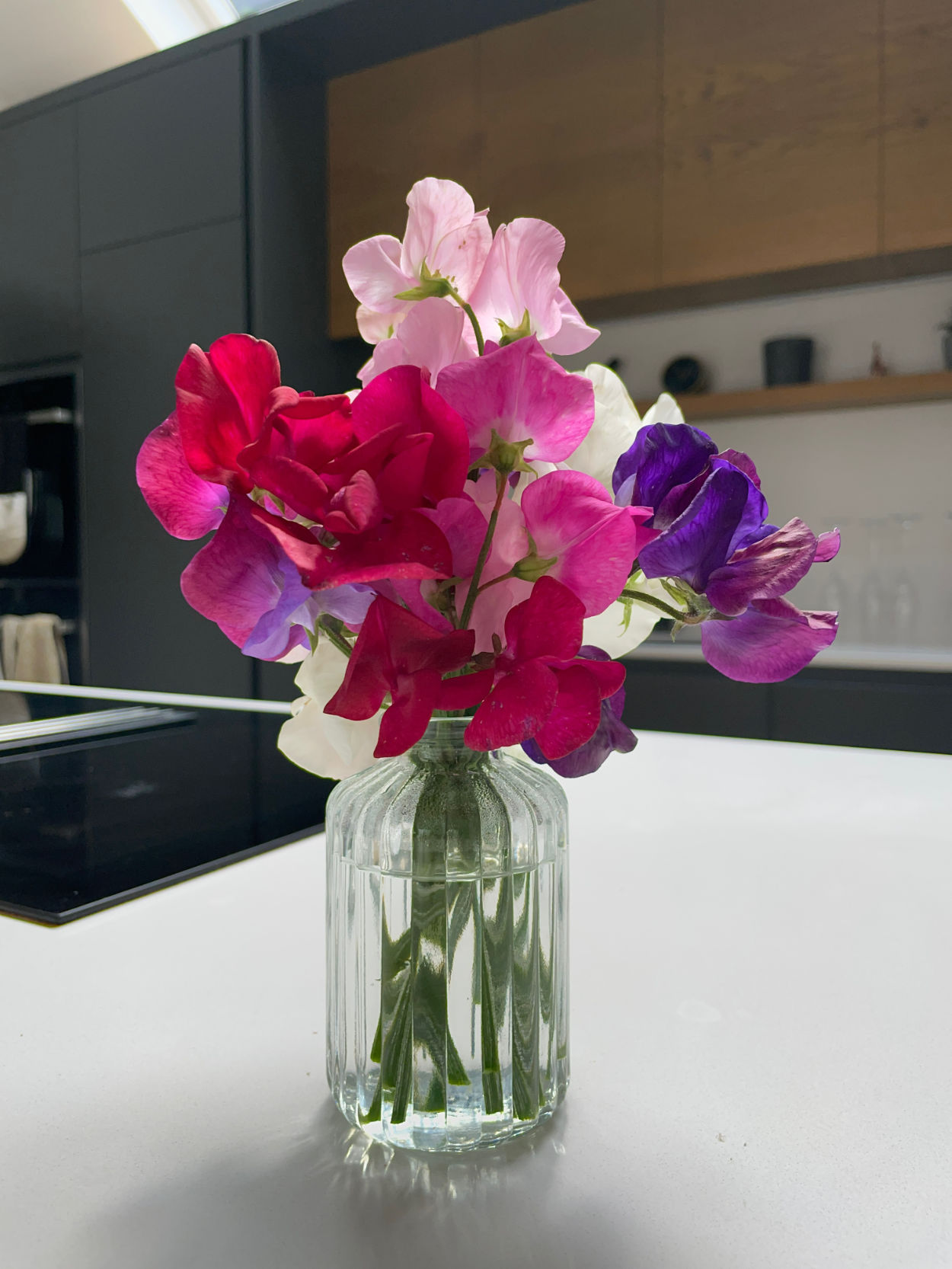 A small bouquet of pink and purple sweet peas in a glass bottle on a white kitchen counter